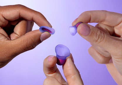 Close-up of three hands holding and demonstrating the flexibility of purple silicone sleep earplugs against a light purple background.